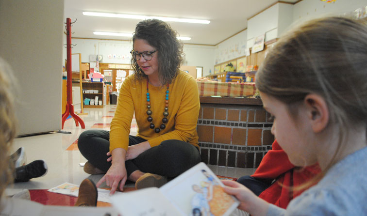 Teacher aide sitting on kindergarten classroom floor reading aloud with students