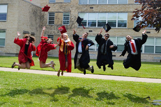 six graduates in red and black gowns jumping in the air and throwing their caps