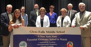 Adults and one student pose behind a banner that reads "Glens Falls Middle School, a New York State Essential Elements: School to Watch"
