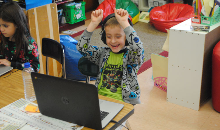 boy in sweatshirt wearing headphones plugged into a Chromebook with fists raised smiling