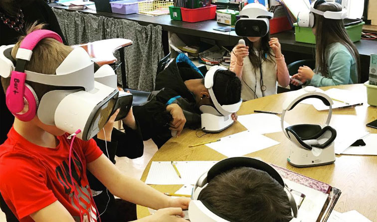 five students sitting at a table with virtual reality headsets covering their eyes and ears