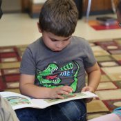 kindergarten student reading book in lap on classroom floor with girl looking on