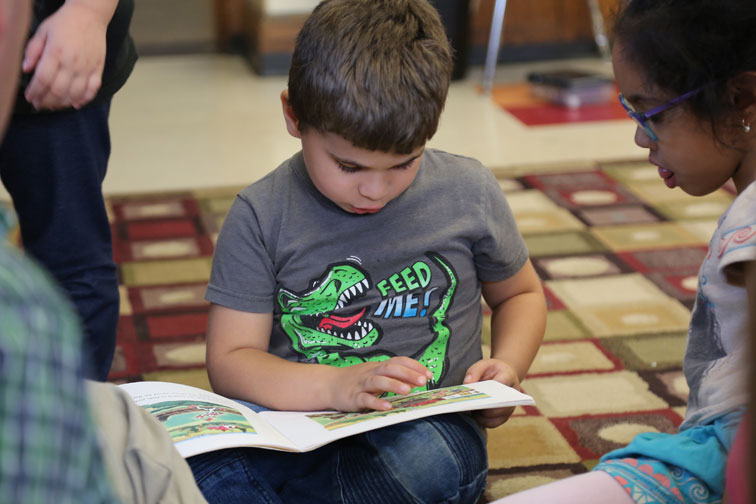 kindergarten student reading book in lap on classroom floor with girl looking on