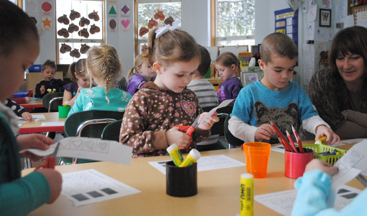 several pre-k students sitting at classroom table cutting paper with glue sticks on table