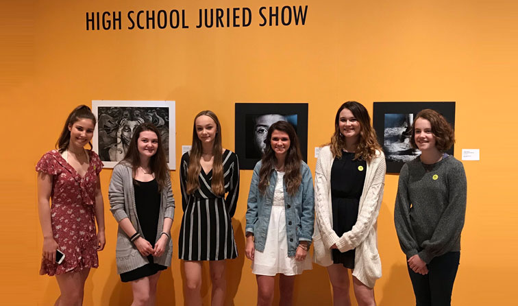 six students smiling in front of orange wall with artwork hanging behind