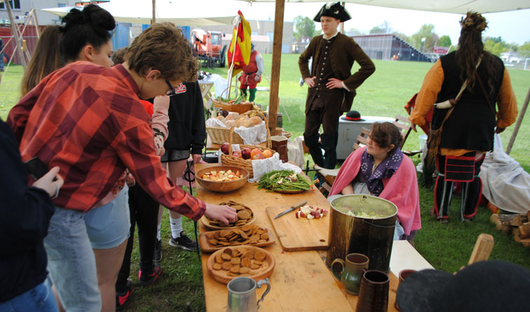 five students and re-enactors around a colinial camp kitchen table with cookies and fresh vegetables