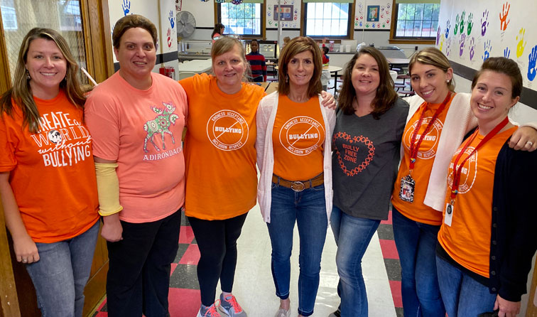 seven people standing in school cafeteria with orange anti-bullying shirts, smiling