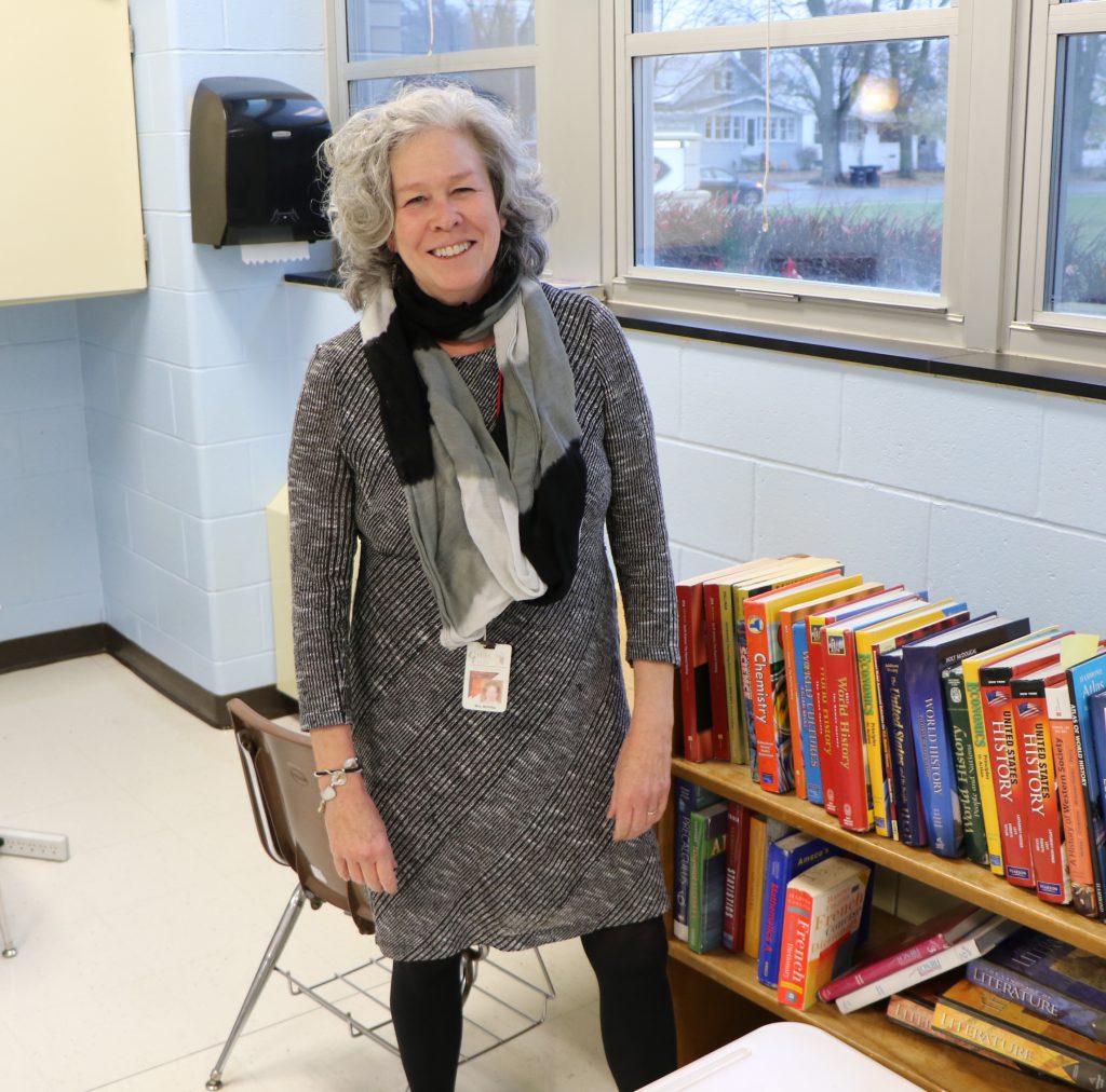 teacher smiling in classroom with stack of books