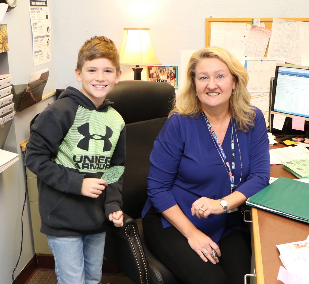student and staff member smiling behind office desk with green turtle hall pass
