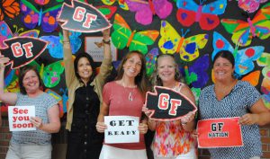 five faculty and staff members holding welcome signs and smiling in front of colorful bulletin board