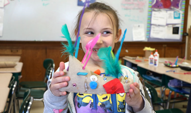 student smiling behind colorful art creation with feathers and cardboard