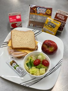 grab and go meal featuring turkey and cheese sandwich, green salad with drerssing, apple, graham crackers, apple juice, cocoa puffs and chocolate milk