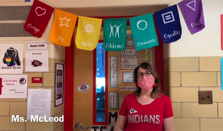 teacher standing in fron t of classroom door wearing mask with colorful banner above reading courage joy spirit celebrate community equality diversity