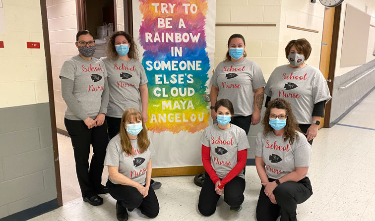 Seven school nurses wearing matching shirts and face masks by a painted sign reading "try to be a rainbow in someone else's cloud - Maya Angelou"