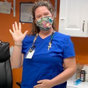 school nurse in blue scrubs and flowered face mask, waving in the school health office