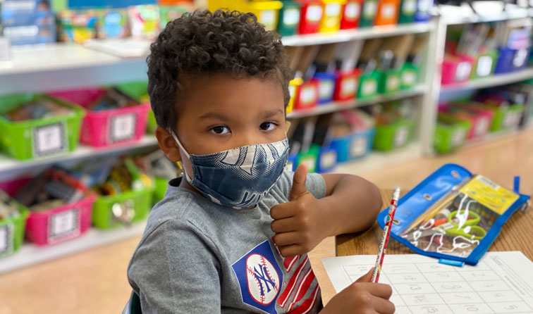 elementary student in front of colorful classroom bins giving thumbs-up with face mask on