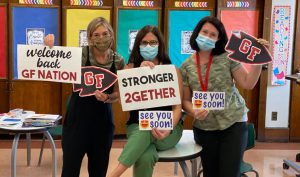 Three teachers in a classroom smiling under their masks and holding welcome back signs