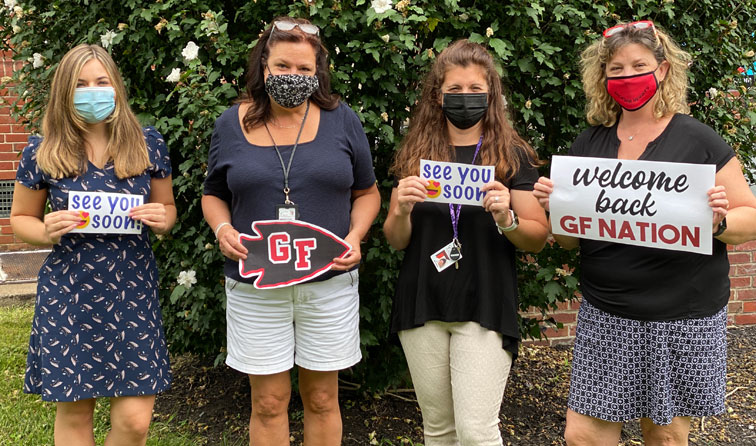 Four teachers outside school smiling under their masks and holding welcome back signs