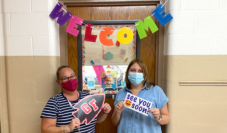 Two teachers in a classroom smiling under their masks and holding welcome back signs