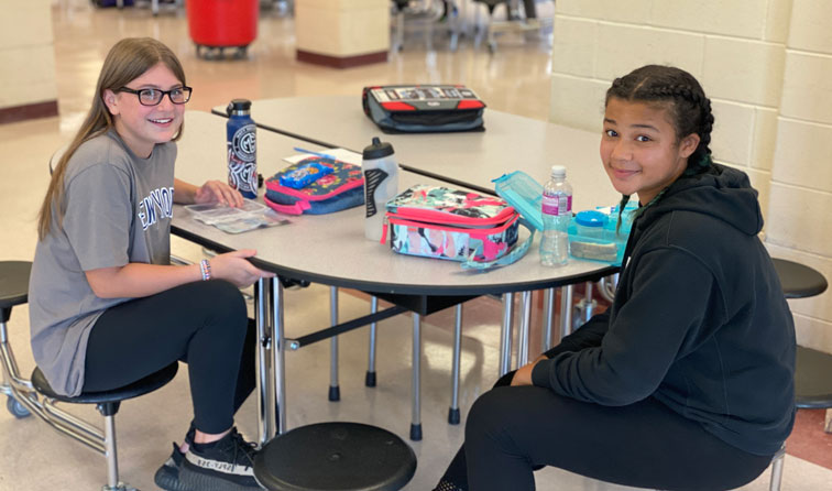two students at an MS lunchroom table with lunchboxes and water bottles on top