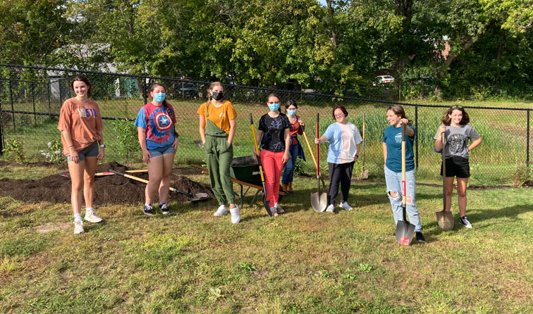 eight students holding shovels standing in front of a fence with shrubs and plants behind them