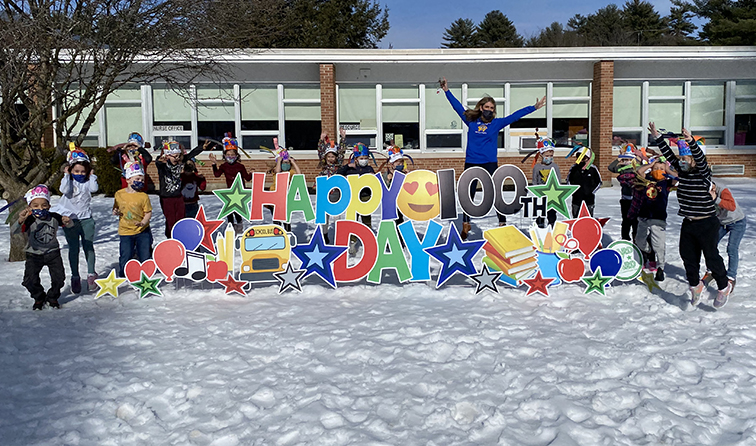 group of students on snowy front lawn of school jumping in mid-air around yard signs that read: happy 100th day of school