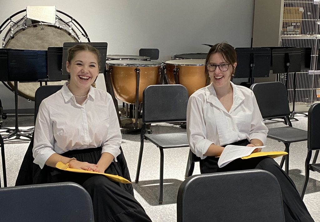 two students in concert dress, sitting in a school band room smiling