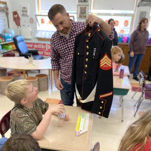 Veteran Marine showing uniform jacket to student seated at desk in classroom