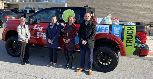 four people smiling in front of a bright red pickup truck from Lia Auto Group that has several huge bags of donated gifts in the back of it