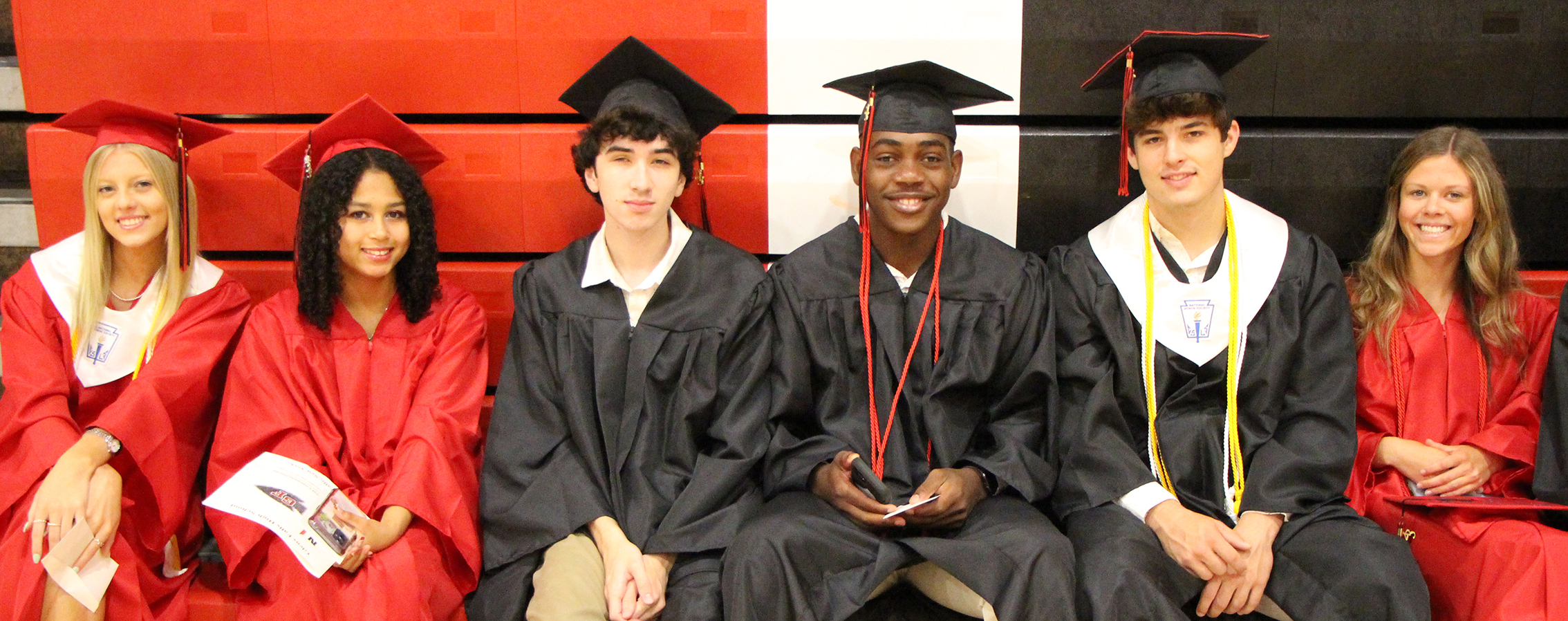 several high school graduates in their red and black caps and gowns prepare for the ceremony in the high school gym